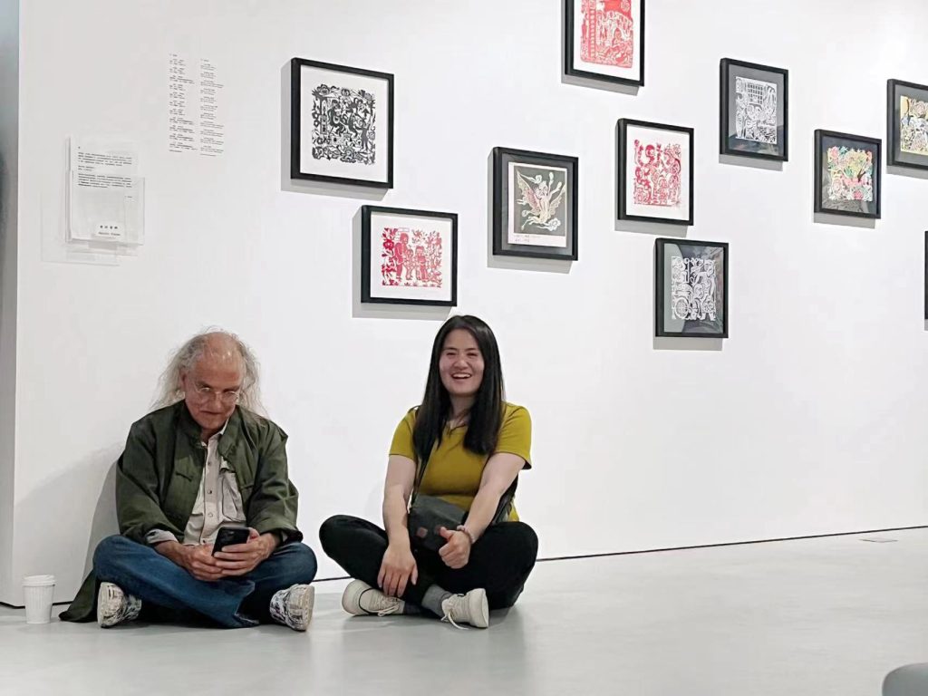 Two figures sit on the floor underneath a museum display of artwork.