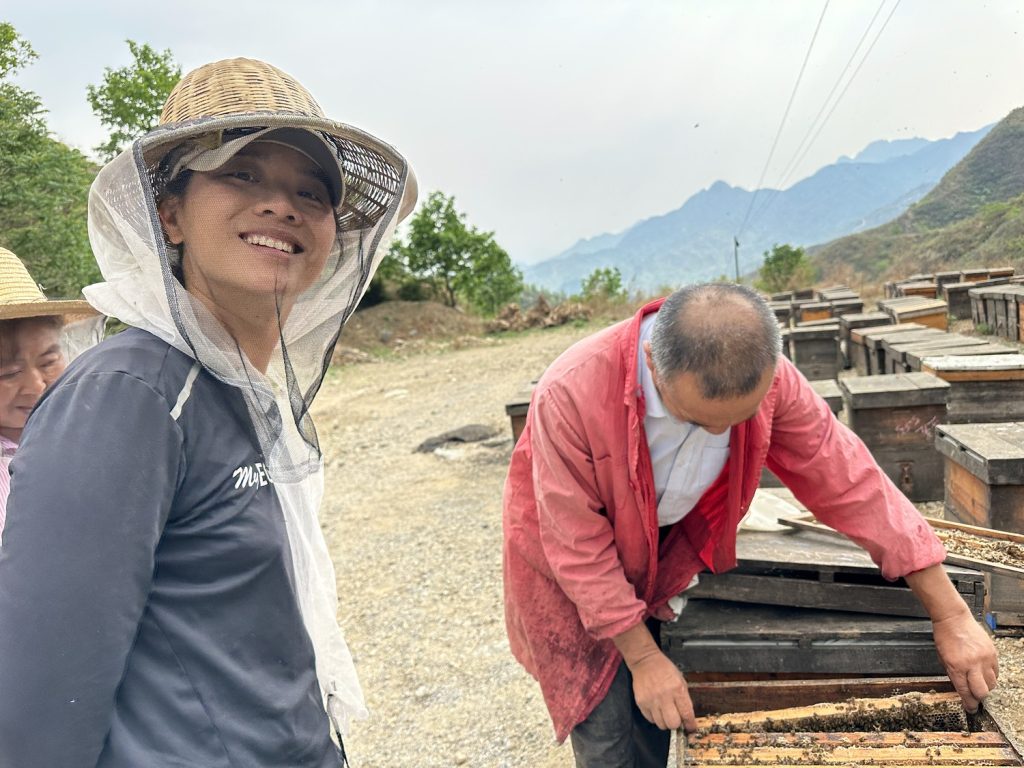 A person in a beekeeping hat smiles as another person adjusts a beekeeping box to the right.