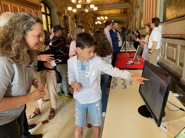 A young student indicates a computer screen to an adult looking on in a crowded hallway in a state capital building.