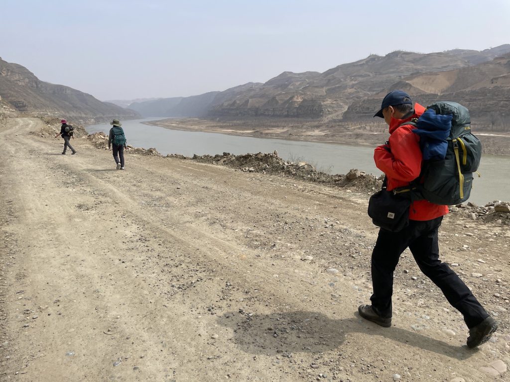 Three people wearing backpacks walk on a dusty trail alongside a low river.